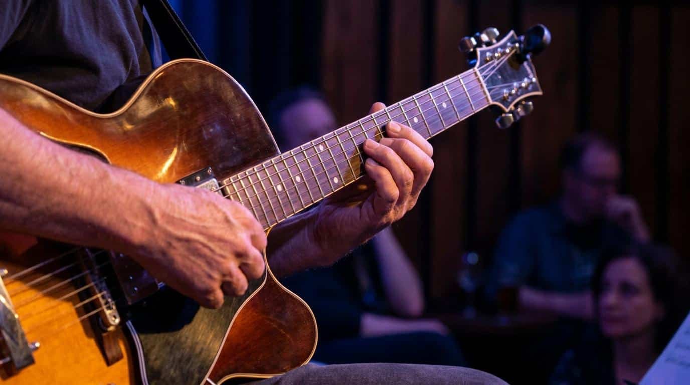 Maîtriser la gamme mineure mélodique en jazz Gros plan sur les mains d'un guitariste jouant sur une guitare jazz archtop dans une salle sombre.
